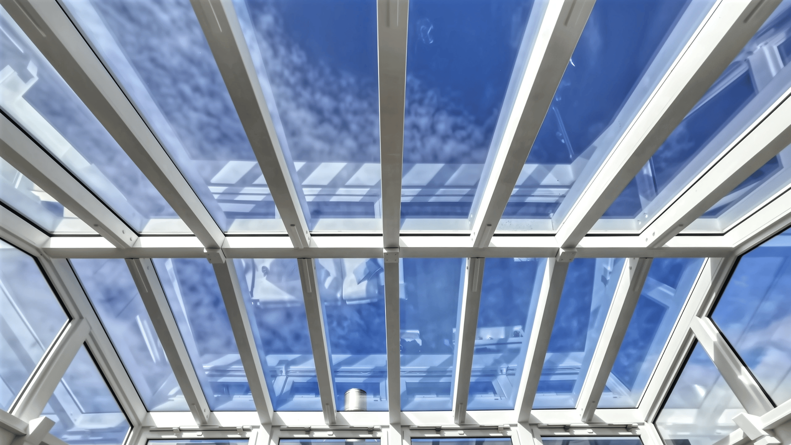 Architectural atrium glass roof structure showing panels, beams, and integrated ventilation windows against blue sky.