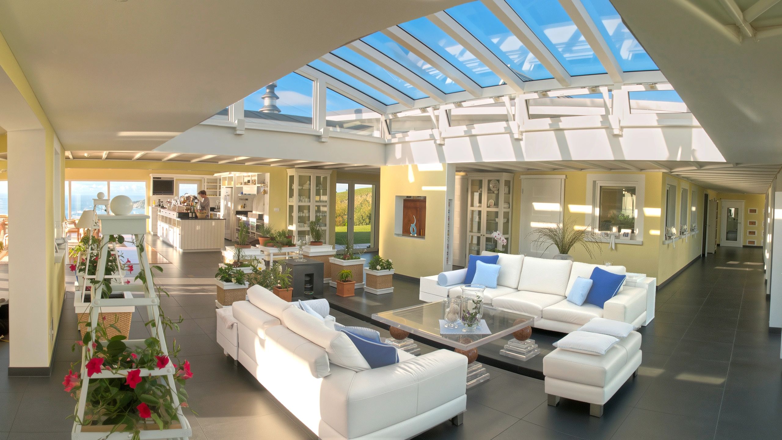 Expansive open floor design in big luxury house featuring glass roof Atrium, white sofas, and view towards kitchen.