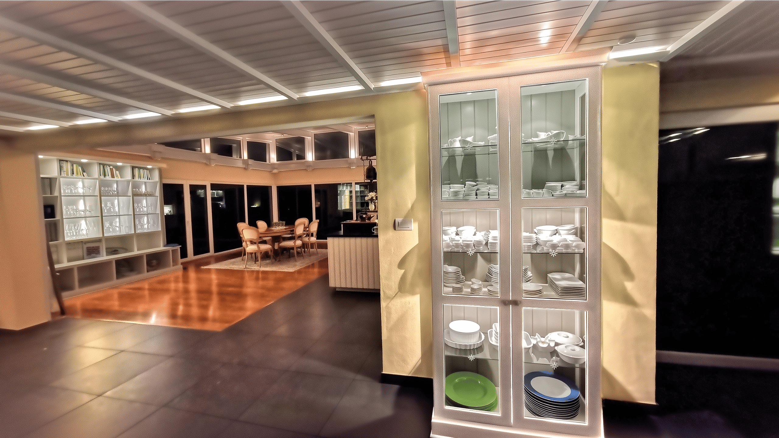 Illuminated white kitchen display cabinet with porcelain near walkway ceiling lighting and background library.