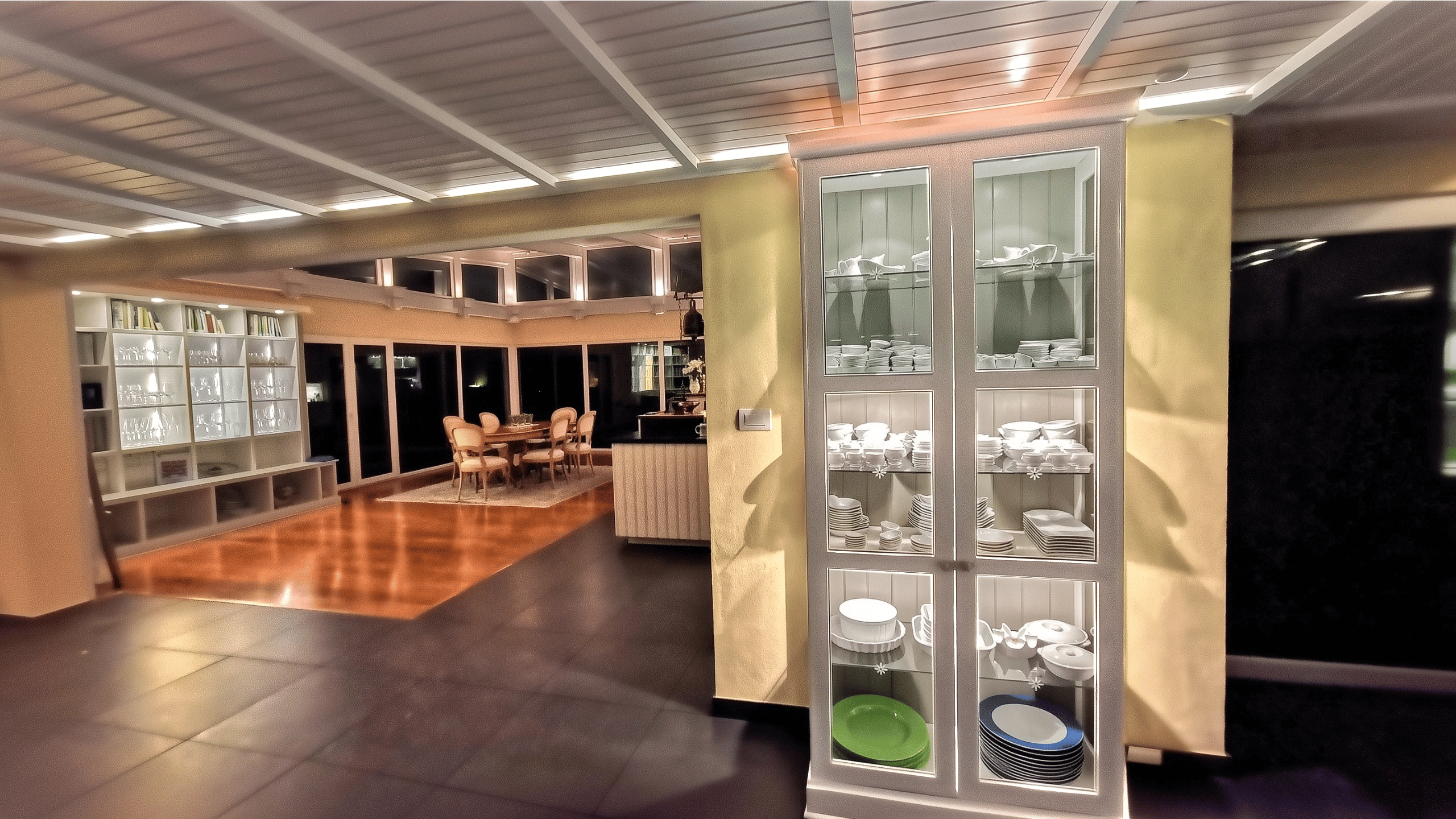 Illuminated white kitchen display cabinet with porcelain near walkway ceiling lighting and background library.